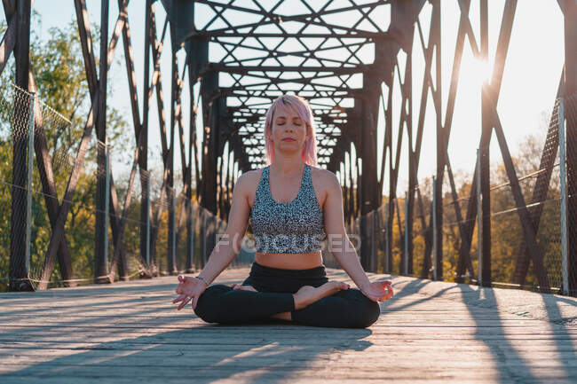 Уровень Ground женщины с закрытыми глазами, сидящей в позе Padmasana во время практики йоги на пешеходной дорожке моста в солнечном свете — стоковое фото