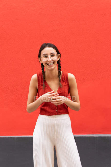 Весела молода жінка в зачісці для кісок, дивлячись на камеру на червоному тлі на вулиці — стокове фото