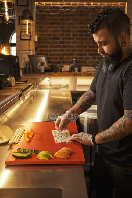 Бічний погляд на чоловічого кухаря в рукавицях і одноманітну підготовку апетитних суші рулони за столом в азіатському ресторані. — Stock Photo