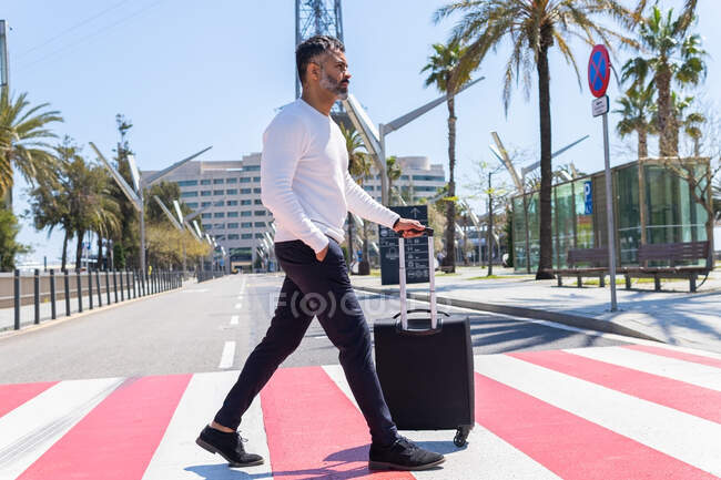 Side view full length serious adult male in smart casual wear strolling with hand in pocket and suitcase across road on modern sunny street — Stock Photo
