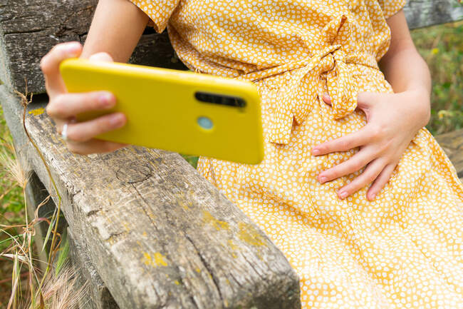 Recadrée femelle enceinte méconnaissable en robe toucher le ventre et prendre l'autoportrait sur téléphone portable tout en étant assis sur le banc à la campagne en été — Photo de stock