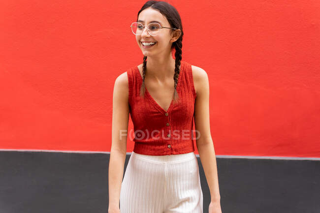 Cheerful young female in pigtails hairstyle while looking away on red background in street — Stock Photo