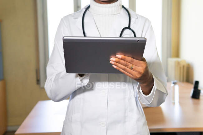 Crop jovem médico feminino preto em casaco médico com estetoscópio trabalhando com tablet no escritório de clínica moderna — Fotografia de Stock