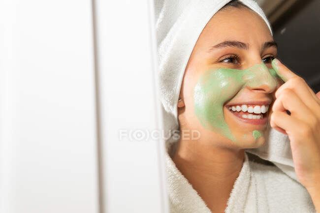 Donna felice con asciugamano sulla testa sorridente e diffusione maschera verde sul viso mentre si guarda specchio in bagno a casa — Foto stock