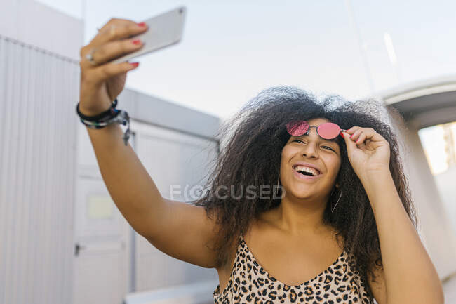 Mujer joven y afro con gafas de sol sonriendo y tomando una selfie con su teléfono inteligente - foto de stock