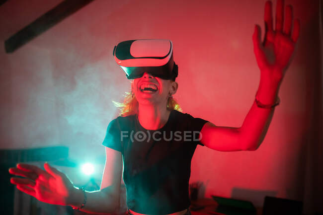Donna felice che indossa t-shirt mentre utilizza occhiali VR e in piedi in studio con luci al neon rosse — Foto stock