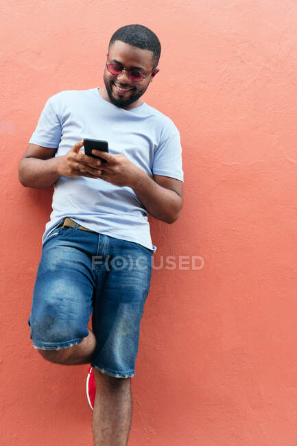 Homme noir souriant regardant son téléphone intelligent tout en se tenant dans la ville — Photo de stock