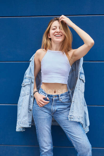 Молодая веселая женщина в джинсовой куртке и джинсах стоит на фоне синей стены на улице и смотрит в камеру — стоковое фото