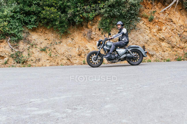 Ganzkörper-bärtiger Biker in schwarzer Lederjacke und Helm auf modernem Motorrad auf asphaltierter Straße inmitten üppig grüner Bäume, die im bergigen Tal wachsen — Stockfoto