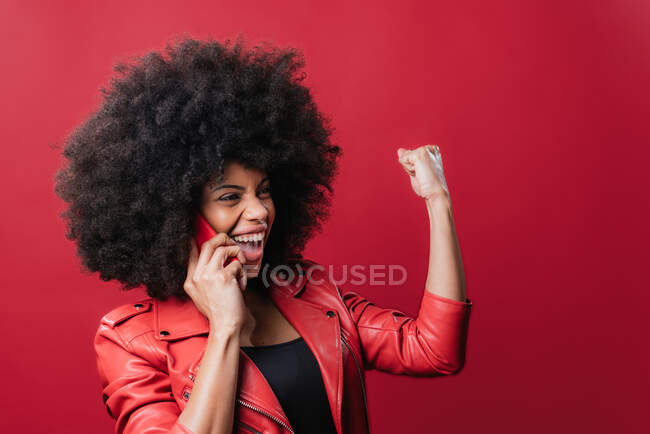 Femme afro-américaine excitée avec poing serré faire appel sur un téléphone mobile et célébrer le succès sur fond rouge — Photo de stock