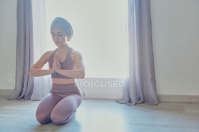 Jovem fêmea concentrada em sportswear com olhos fechados e mãos namaste sentado no tapete de ioga em casa em volta iluminado — Fotografia de Stock