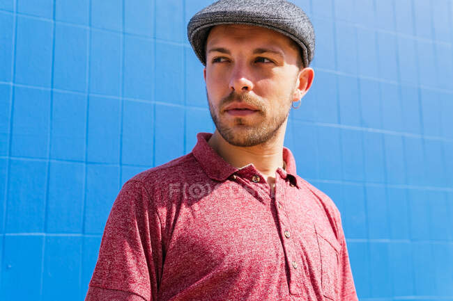 Продуманий молодий бородатий чоловік у стильному повсякденному одязі та кепці насолоджується літнім днем біля синьої стіни на міській вулиці — стокове фото