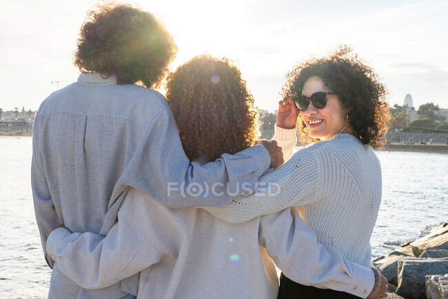 Задний вид анонимных друзей с вьющимися волосами, стоящих близко в объятиях против городского пейзажа и моря в солнечном свете — стоковое фото