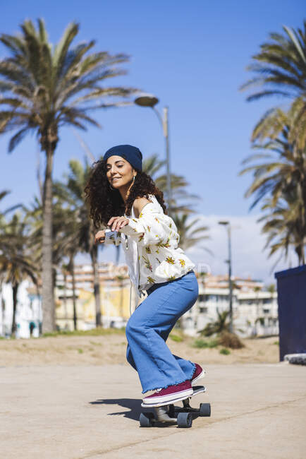 Все тело активной счастливой женщины в повседневной одежде катается на скейтборде по дороге вдоль песчаного пляжа и высокие пальмы во время тренировки — стоковое фото