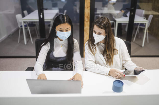 Багаторасові жінки-колеги в масках сидять за столом з ноутбуком і обговорюють бізнес-проект під час роботи в коворкінгу — стокове фото