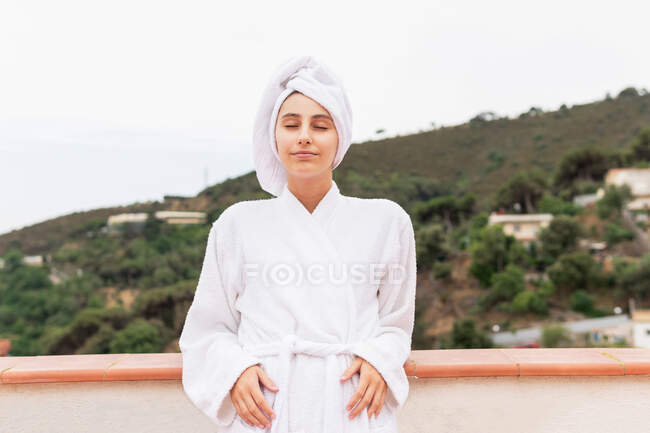 Ottimista giovane donna in accappatoio e asciugamano sorridente mentre si rilassa sul balcone durante la routine di cura della pelle nel fine settimana — Foto stock