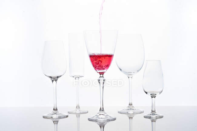 Anonymous person pouring red sparkling wine into goblet laced on mirrored table near various shaped glasses against white background — Stock Photo