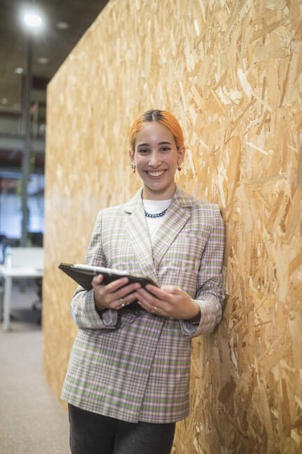 Улыбающаяся предпринимательница, стоящая с компьютером у стены в коворкинге, глядя в камеру — стоковое фото