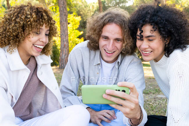 Groupe de femmes gaies et l'homme aux cheveux bouclés assis sur plaid coloré sur la pelouse dans un parc ensoleillé et regarder smartphone ensemble — Photo de stock