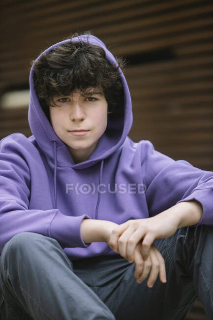Adolescente em roupas casuais com capuz olhando para a câmera enquanto sentado no skate na parede na rua com folhas caídas — Fotografia de Stock