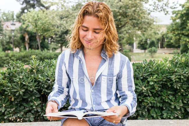 Fröhlicher Mann mit langen Haaren sitzt am Wochenende auf Bank im Garten und liest interessante Geschichte in Buch — Stockfoto