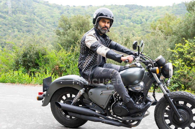 Full body of focused bearded ethnic male biker in black leather jacket and helmet riding modern motorbike on asphalt road amidst lush green trees growing in mountainous valley — Stock Photo