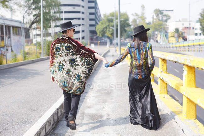 Обратный вид на неузнаваемую стильную пару в хипстерской одежде, держащуюся за руки и идущую по улице летом — стоковое фото