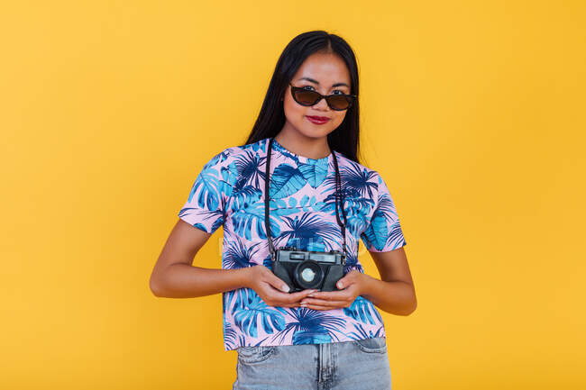 Happy Asian female in t shirt with tropical leaf print holding photo camera on yellow background in studio — Stock Photo