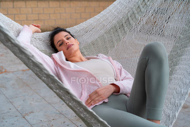 Молода жінка в спортивному одязі лежить в гамаку над плиткою прогулянки, дивлячись на камеру вдень — стокове фото