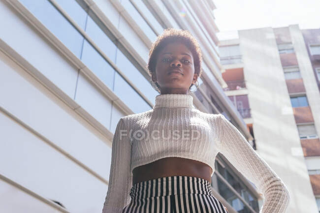 Von unten stylische selbstbewusste Afroamerikanerin, die die Hand auf der Taille hält und in die Kamera blickt, während sie an sonnigen Tagen in der Stadt in der Nähe moderner Wohnhäuser steht — Stockfoto