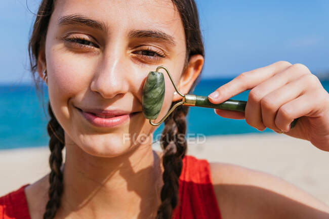Содержание женщины с помощью нефритового ролика и делать массаж лица, глядя на камеру на берегу моря летом — стоковое фото