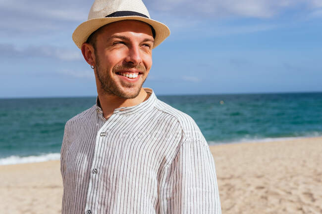 Молодой бородатый мужчина в модной повседневной одежде и кепке стоит, глядя на песчаный пляж в сторону моря, проводя летние каникулы на берегу моря — стоковое фото
