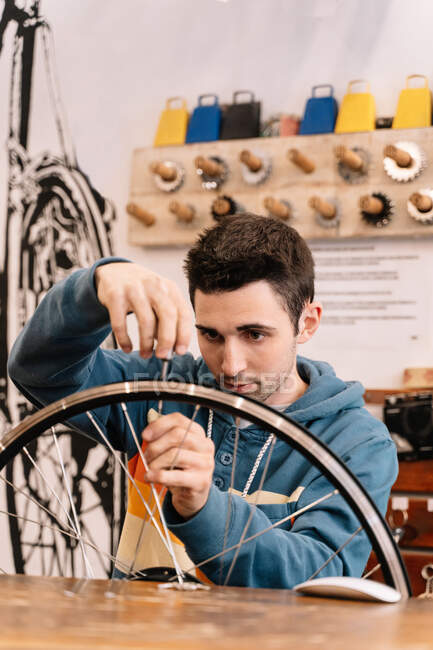 Serious male mechanic fixing wheel spoke while sitting at workbench in bike service — Stock Photo