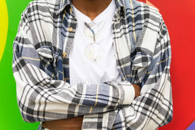 Crop unkenntlich ethnischen männlichen Hipster im trendigen karierten Hemd und mit Brille stehen mit verschränkten Armen auf buntem Hintergrund in der Stadt — Stockfoto