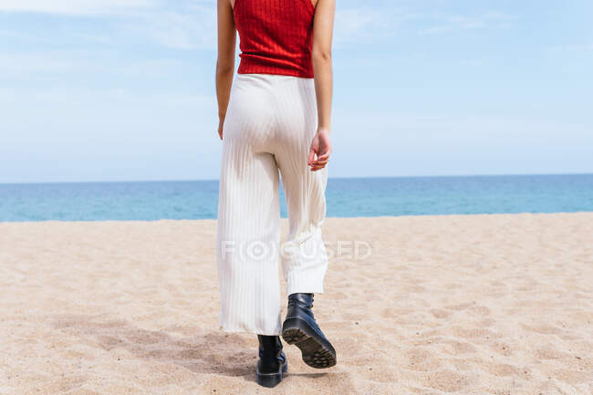 Rückansicht einer anonymen Frau in Stiefeln, die an einem sonnigen Tag am Sandstrand in Richtung ruhiges blaues Meer läuft — Stockfoto