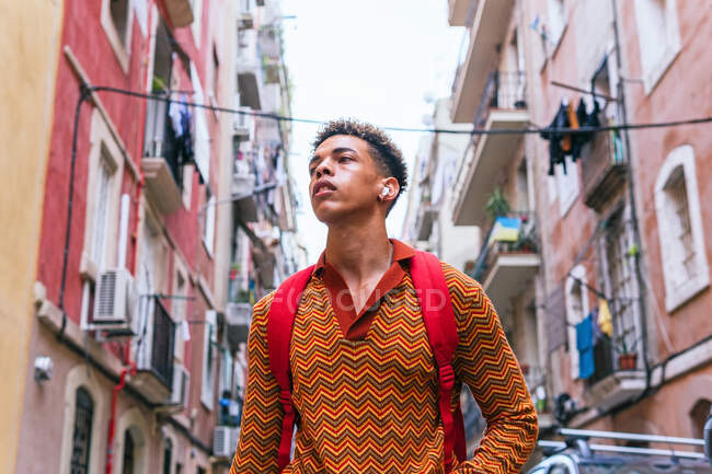 Low angle of confident young ethnic male tourist with backpack and TWS earbuds looking away while exploring old narrow streets of Barcelona city — Stock Photo