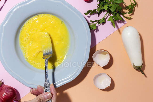 Visão superior da cultura fêmea irreconhecível batendo ovo não cozido contra raminhos de salsa fresca e berinjela branca em fundo de duas cores — Fotografia de Stock
