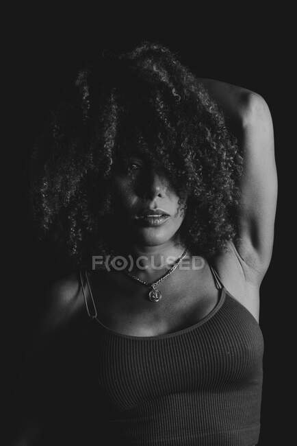 Черно-белая очаровательная афроамериканская модель с кудрявыми волосами смотрит на камеру в темной студии — стоковое фото