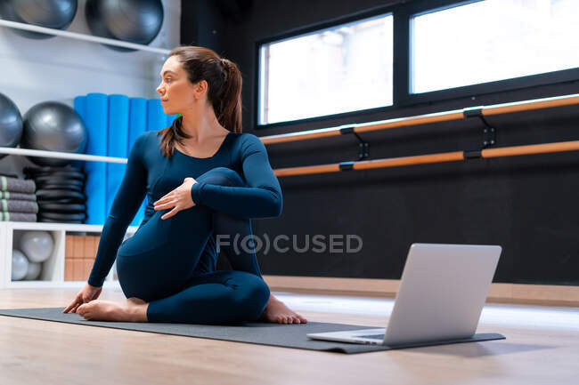 Cuerpo completo de instructora de yoga joven y flexible haciendo la pose de mitad señor de los peces frente a la pantalla del portátil durante la clase en línea en el gimnasio - foto de stock