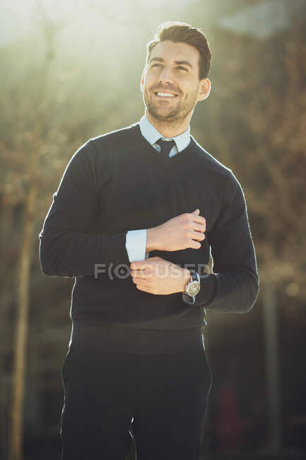Imprenditore maschio barbuto sorridente in orologio da polso con taglio di capelli moderno guardando altrove in città in retroilluminazione — Foto stock