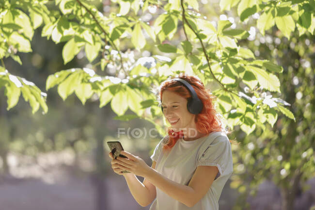 Содержание женщины с рыжими волосами и в наушниках, слушая музыку и просматривая смартфон в парке в солнечный день — стоковое фото
