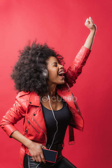 Mulher afro-americana encantada ouvindo música em fones de ouvido e usando telefone celular enquanto dança em fundo vermelho em estúdio — Fotografia de Stock