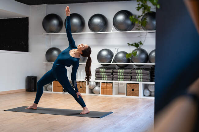 Vista lateral de cuerpo completo de una joven hembra flexible de pie en la pose Exalted Crescent Lunge mientras practica yoga en un moderno gimnasio - foto de stock