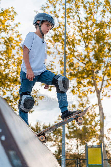 Vista lateral de ángulo bajo del valiente patinador adolescente de pie en el monopatín y preparándose para mostrar truco en la rampa en el parque de skate - foto de stock