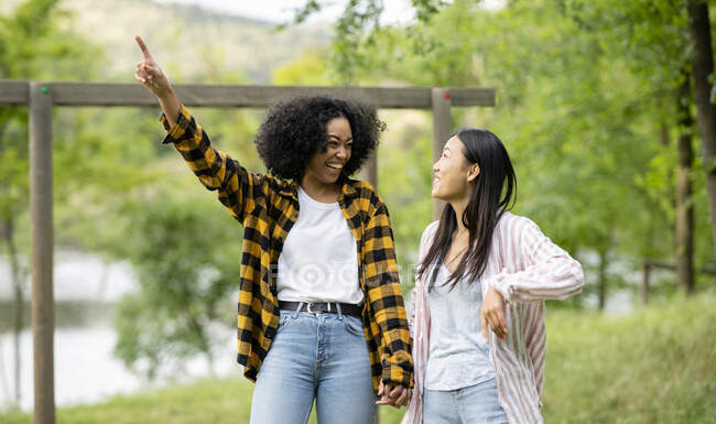 Content LBGT couple of multiracial females holding hands and walking along lake in woods while pointing away — Stock Photo