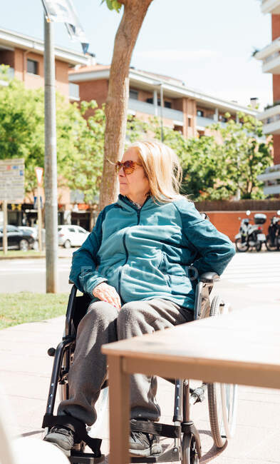 Pensive aged female sitting in wheelchair on street on sunny day and looking away — Stock Photo