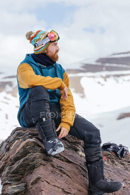Atleta masculino barbudo con ropa deportiva mirando hacia otro lado mientras está sentado en el monte en la temporada de invierno en España - foto de stock
