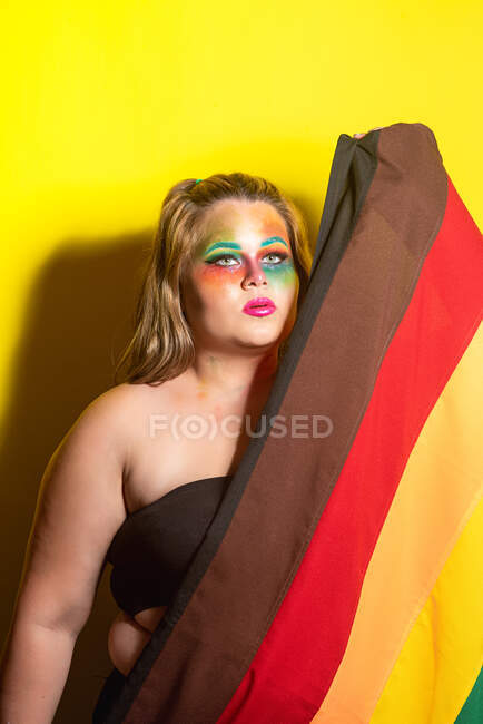 Избыточный вес женская модель с творческим макияжем показывая ЛГБТ-флаг и глядя в сторону на желтый фон — стоковое фото