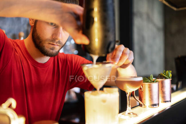 Cantinero enfocado vertiendo cóctel refrescante frío a través del colador en vidrio colocado en el mostrador en el bar - foto de stock
