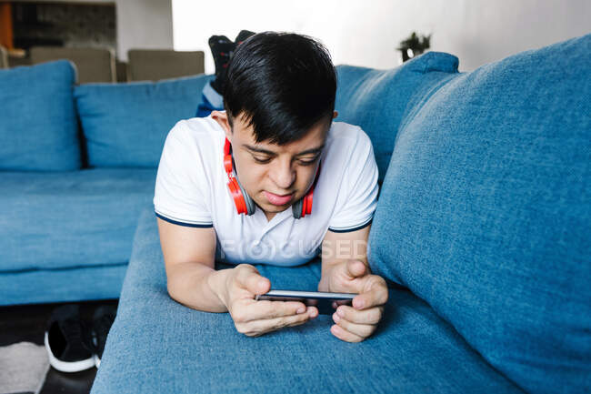 Фокус латинский мальчик, играющий в видеоигры на мобильном телефоне, лежа на диване дома — стоковое фото